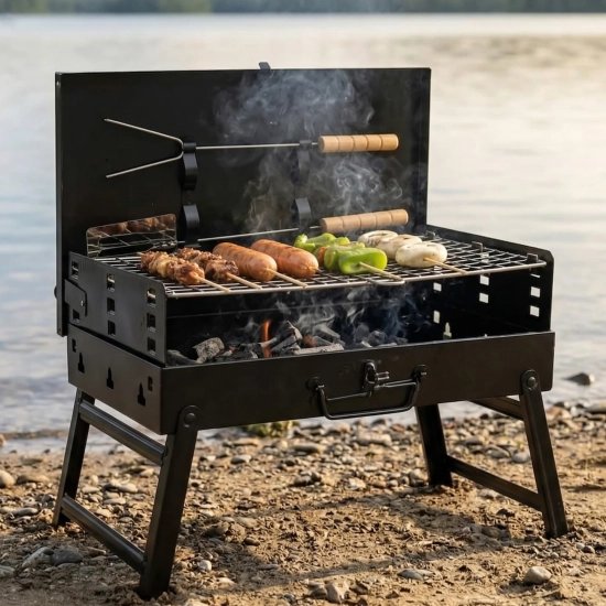 Rozłożony czarny grill przenośny węglowy na kamienistej plaży. Mały grill turystyczny z rusztem, na którym pieką się kiełbaski i szaszłyki podczas plenerowego BBQ