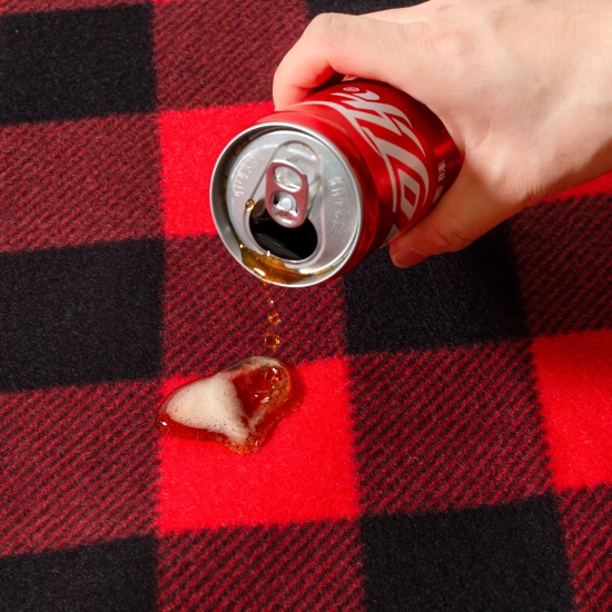 Ciemny napój wylewany z puszki, który perli się na polarowej powierzchni koca w czerwoną kratkę