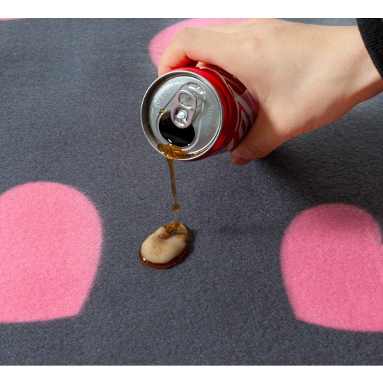 Ciemny napój wylewany z puszki, który perli się na szarej polarowej powierzchni koca w różowe serduszka.