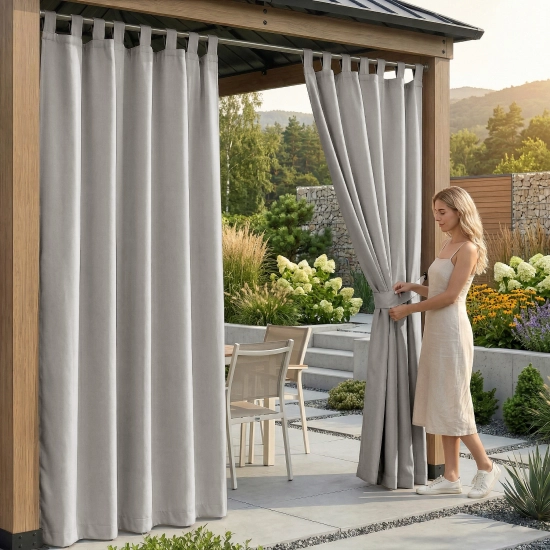 Kobieta pod drewnianą pergolą upinająca jasnoszarą, wodoodporną zasłonę ogrodową na szelkach materiałowym paskiem